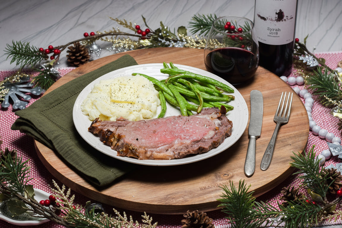 Christmas-themed dinner setup with prime rib, green beans, mashed potatoes, and a glass of red wine on a wooden board.