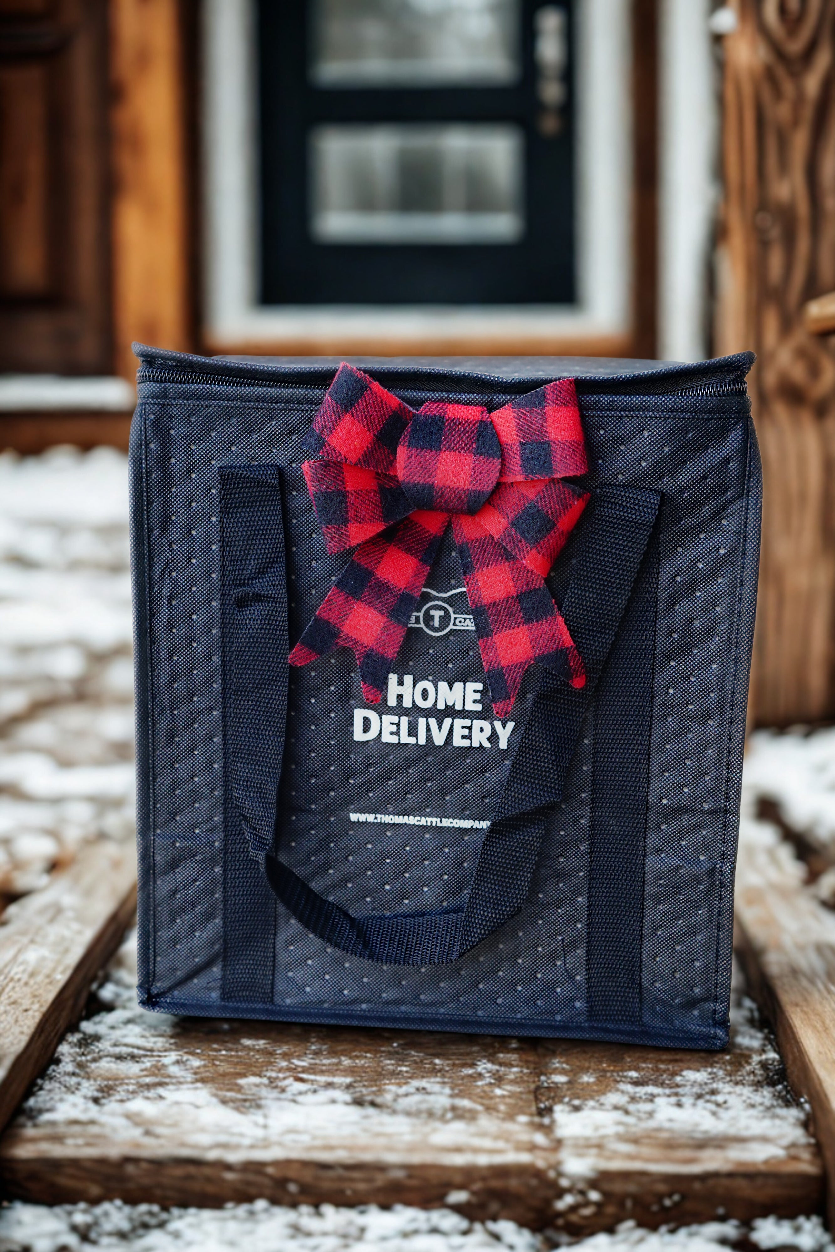 Black delivery bag with red plaid bow and 'Home Delivery' text on a snowy surface.